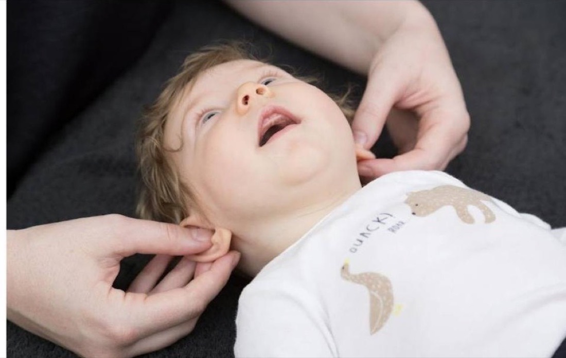 A baby lying on a dark blanket looking upward, with a practitioner's fingertips resting either side of the neck just below the ears.