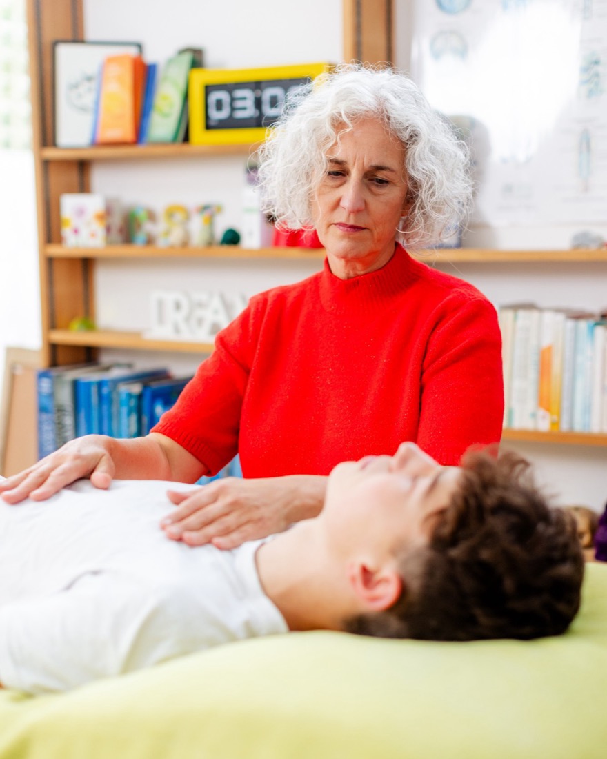 Caroline, in a red jumper, working with both hands resting on the chest of a person lying on the treatment table with their eyes closed. Behind them shelves of books and a chart of the craniosacral system.