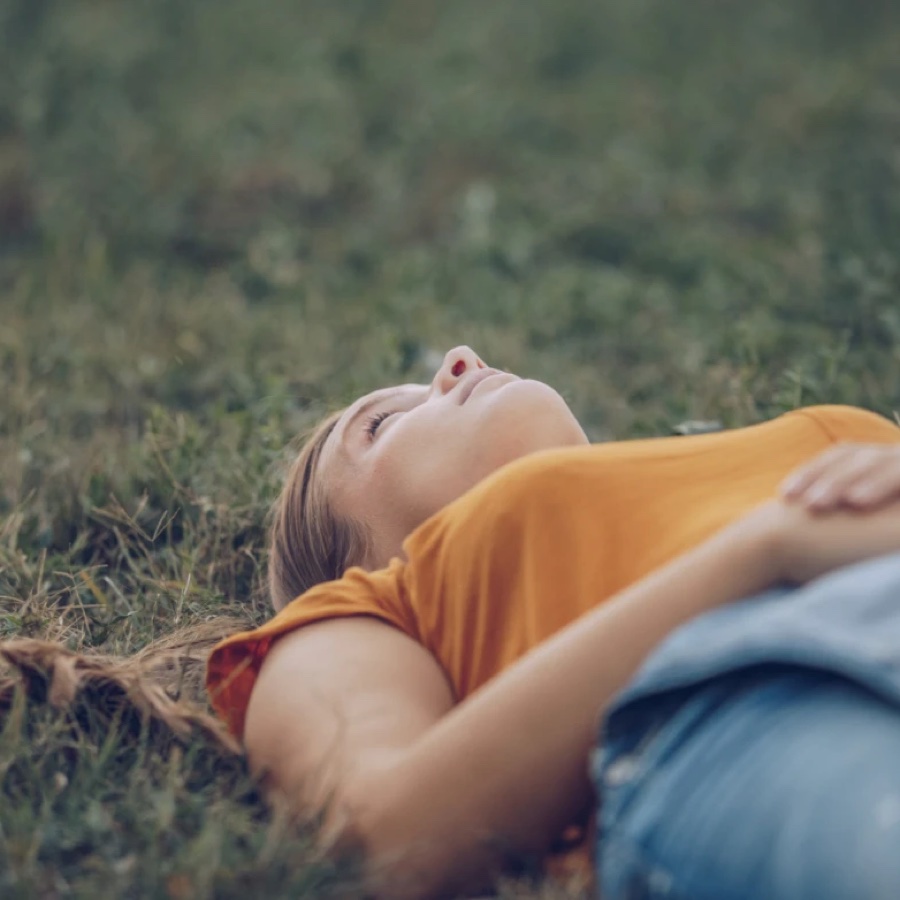 A woman lying on her back on the grass with her eyes closed and one arm across her, her face turned up to the sky.