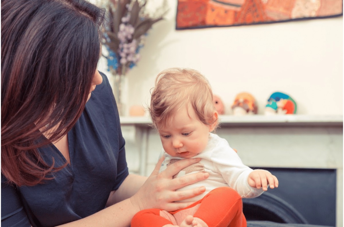 A woman with dark hair sitting with a baby on her lap, one hand resting across the baby's chest.