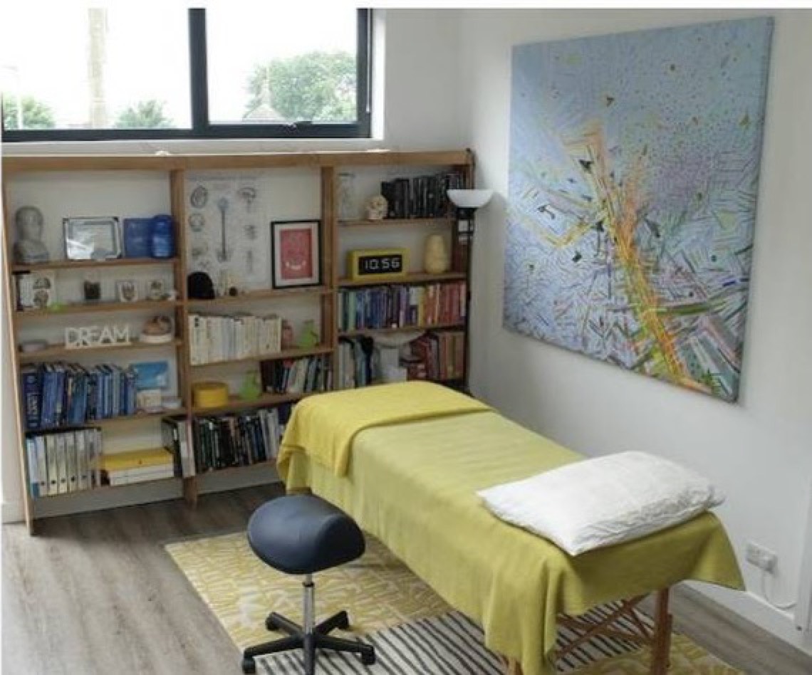 The treatment room: a table draped in yellow with a white pillow and a rolling stool beside it, a wall of bookshelves holding anatomical models and a skull, and a large colourful map of Brighton on the wall.