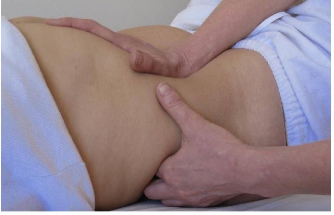 Two hands working gently at a client's lower abdomen, the client lying draped in a pale blue towel.